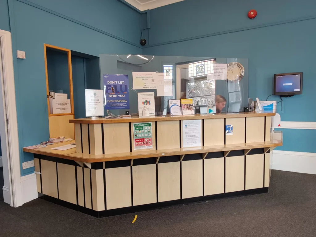 Reception desk at Jesmond Health Partnership with safety screens and informational posters.