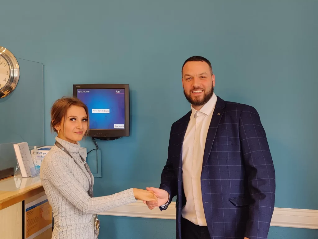 Two professionals shaking hands in a healthcare setting with a blue wall.