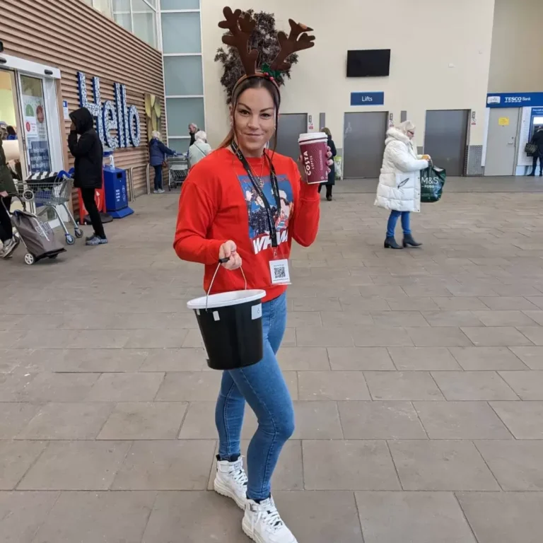 Woman volunteering at Christmas event holding donation bucket and hot drink.