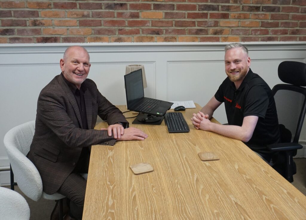 Two men sitting at a desk in an office, smiling, with a computer and brick wall background.